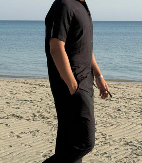 Person in black outfit walking on a sandy beach with ocean and sky in the background