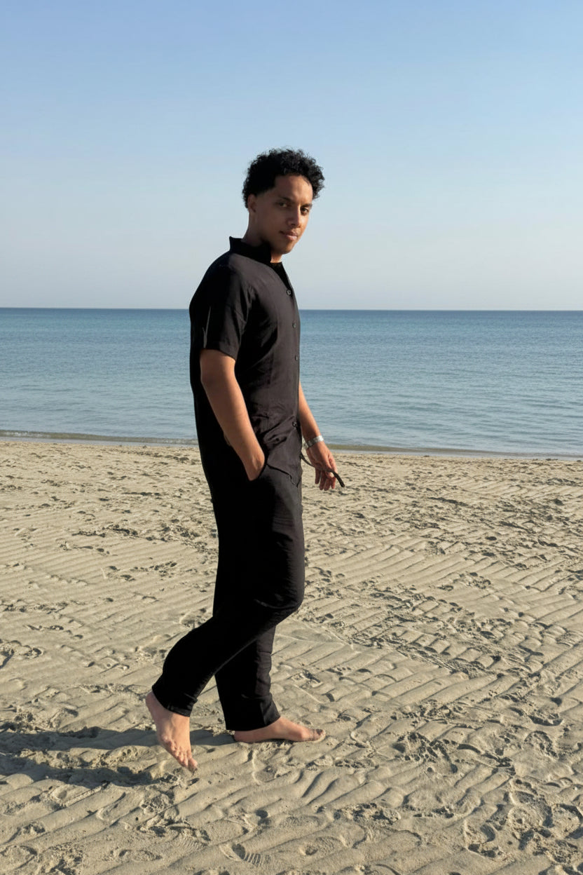 Person in black outfit walking on a sandy beach with ocean and sky in the background